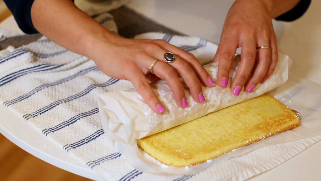 Hands rolling a cake on parchment paper over a striped towel, with pink nail polish and a ring visible.