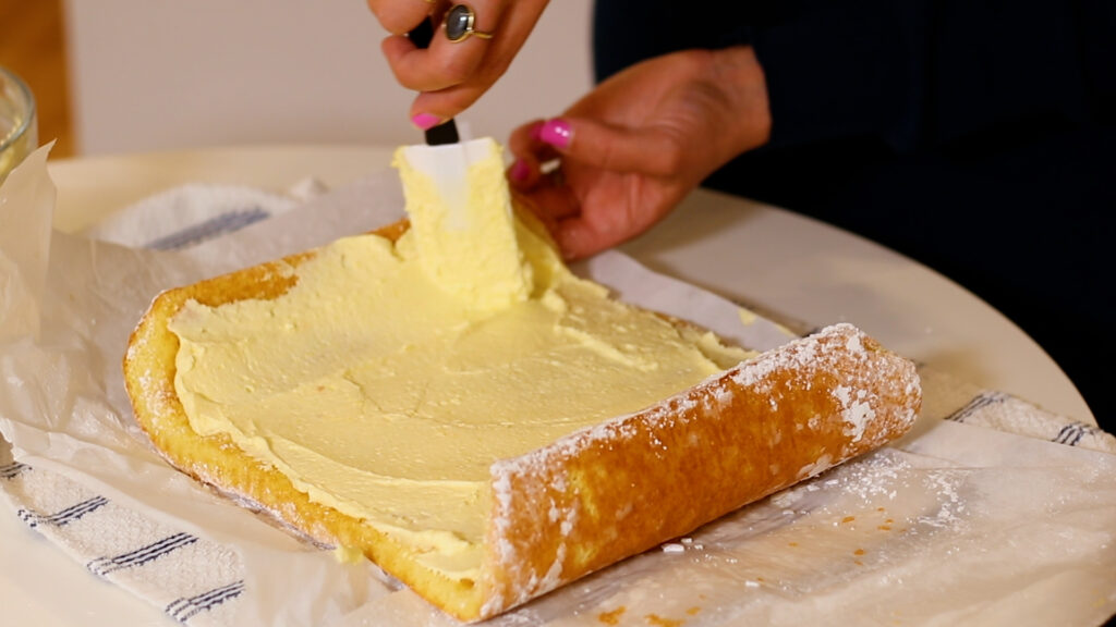 Person spreading cream on a sponge cake sheet with a spatula on a table.