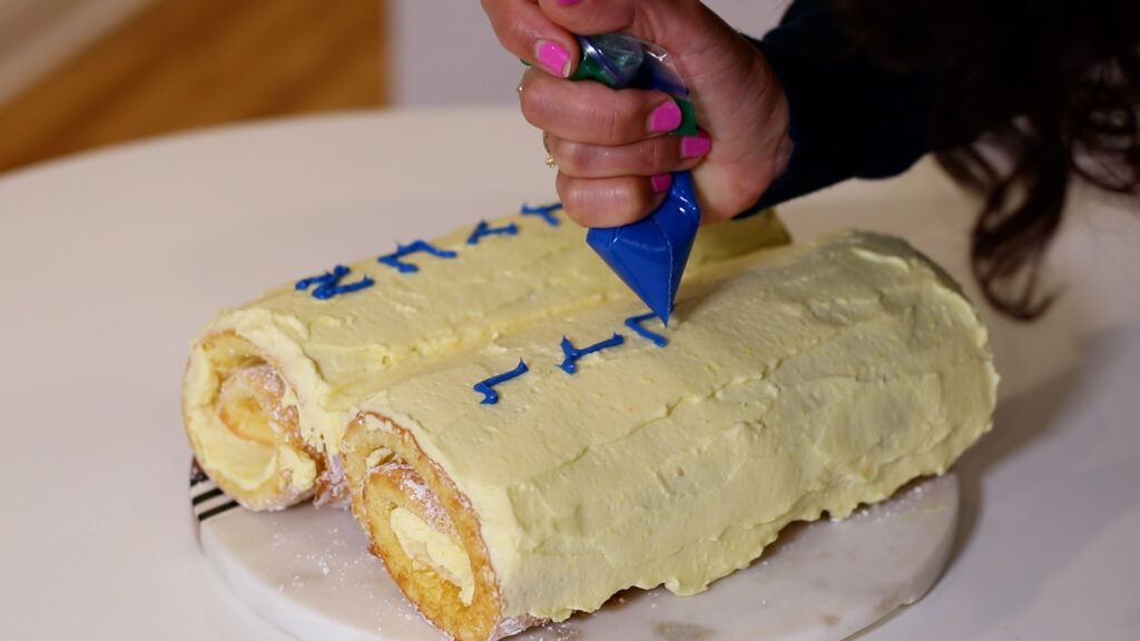 A person decorates a yellow frosted cake with blue icing on a white table.