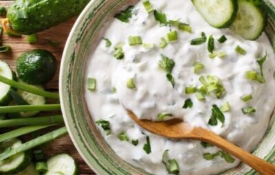 A bowl of creamy tzatziki sauce with cucumber slices and green onions, surrounded by fresh cucumbers on a wooden table.