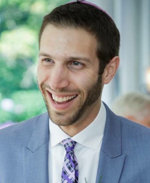 A man in a blue suit and patterned tie smiles warmly indoors.