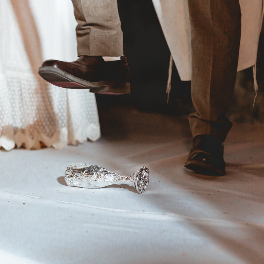 A person in brown shoes stomps on a silver foil-wrapped glass, with a white fabric background.