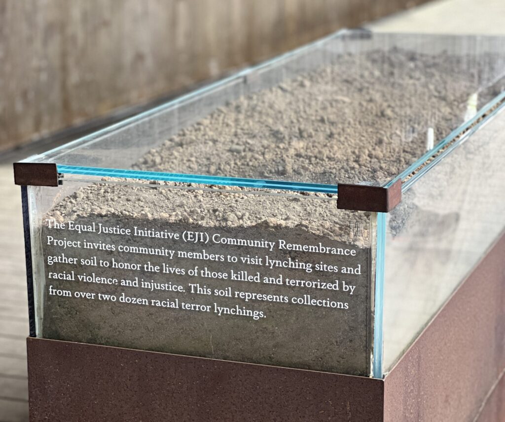 Glass case with soil at EJI display, honoring victims of racial violence and injustice. Text explains its significance.
