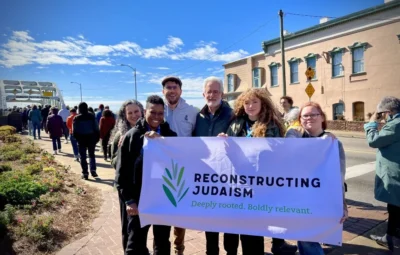Group holding a Reconstructing Judaism banner on a sunny street with buildings and a bridge in the background.