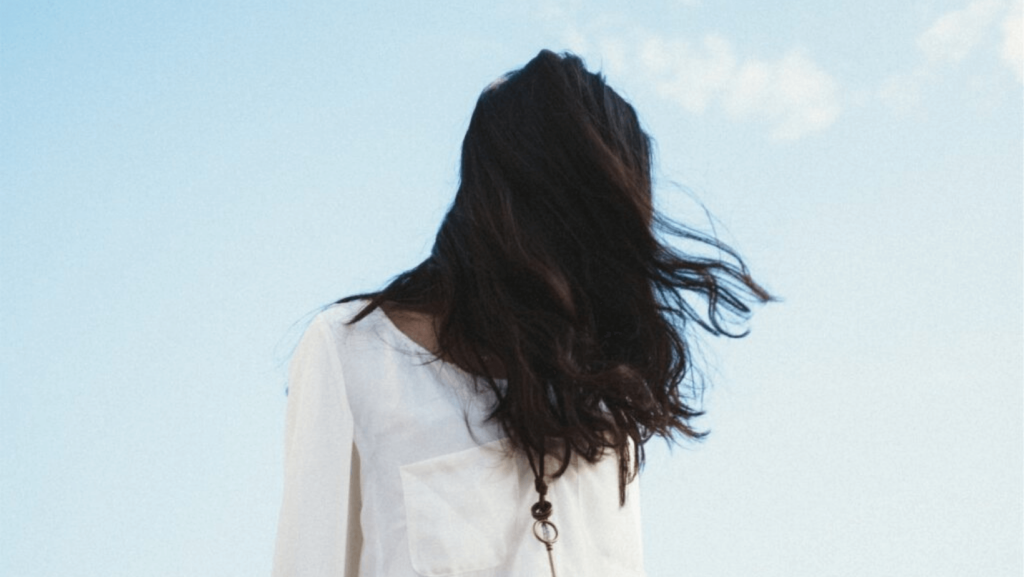 A person in a white shirt standing against a blue sky with long brown hair covering their face