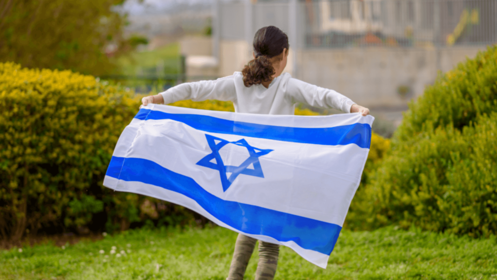 A child with dark curly hair standing on a lawn holds an Israeli flag