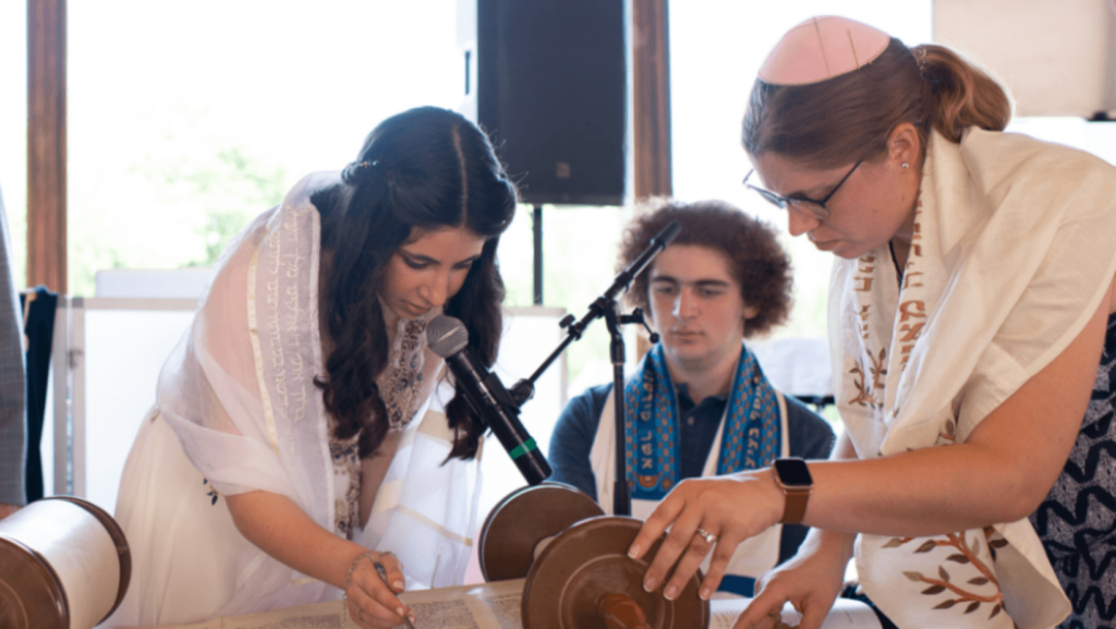 Two young women reading the Torah, a young man sits in the background
