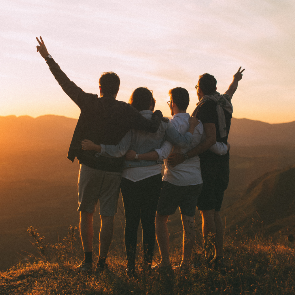 Four people standing on a mountain, embracing and raising arms, watching a sunset.
