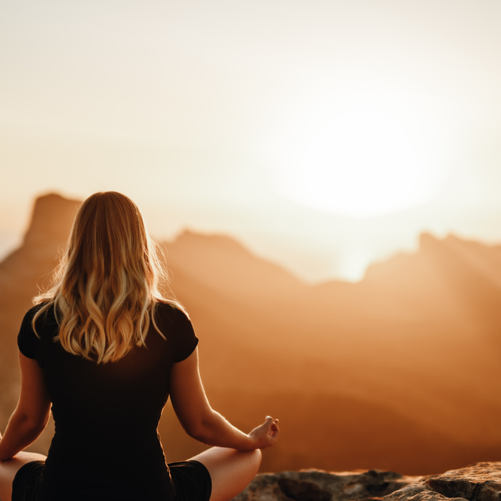 Woman meditating on a mountain top at sunrise, surrounded by a warm, glowing landscape.