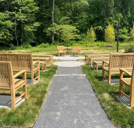 Outdoor seating area with wooden benches and chairs on a grassy area, surrounded by trees.