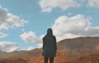 Person in a hooded jacket facing a mountainous landscape under a cloudy blue sky.
