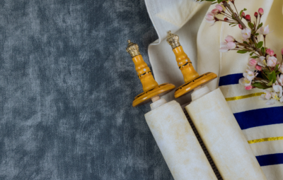 Torah scroll with wooden handles, tallit, and pink flowers on a textured gray background.