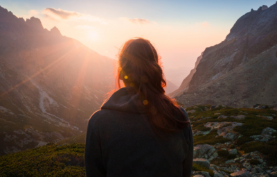 A person sits on a hillside watching a sunrise over distant mountains with a clear sky.