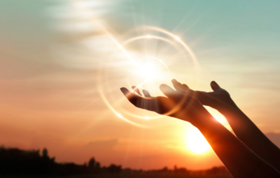 Hands reaching towards a glowing orb in the sky, set against a sunset backdrop.