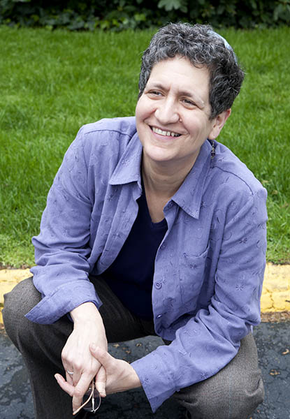 Smiling person with short hair in a blue shirt kneels on the pavement with grass in the background.