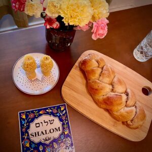 Challah bread on a wooden board, flowers in a vase, candles, and a greeting card saying Shalom on a table.