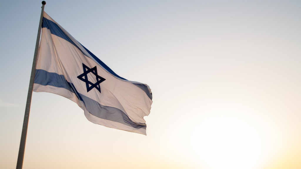 Israeli flag waving against a clear sky with the sun setting in the background.