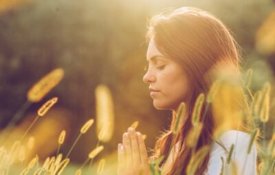 Woman meditating in a sunlit field, surrounded by tall grasses and golden light. Eyes closed, hands together in peace.