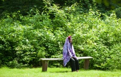 Person sitting on a bench in a lush green park, wrapped in a colorful blanket.