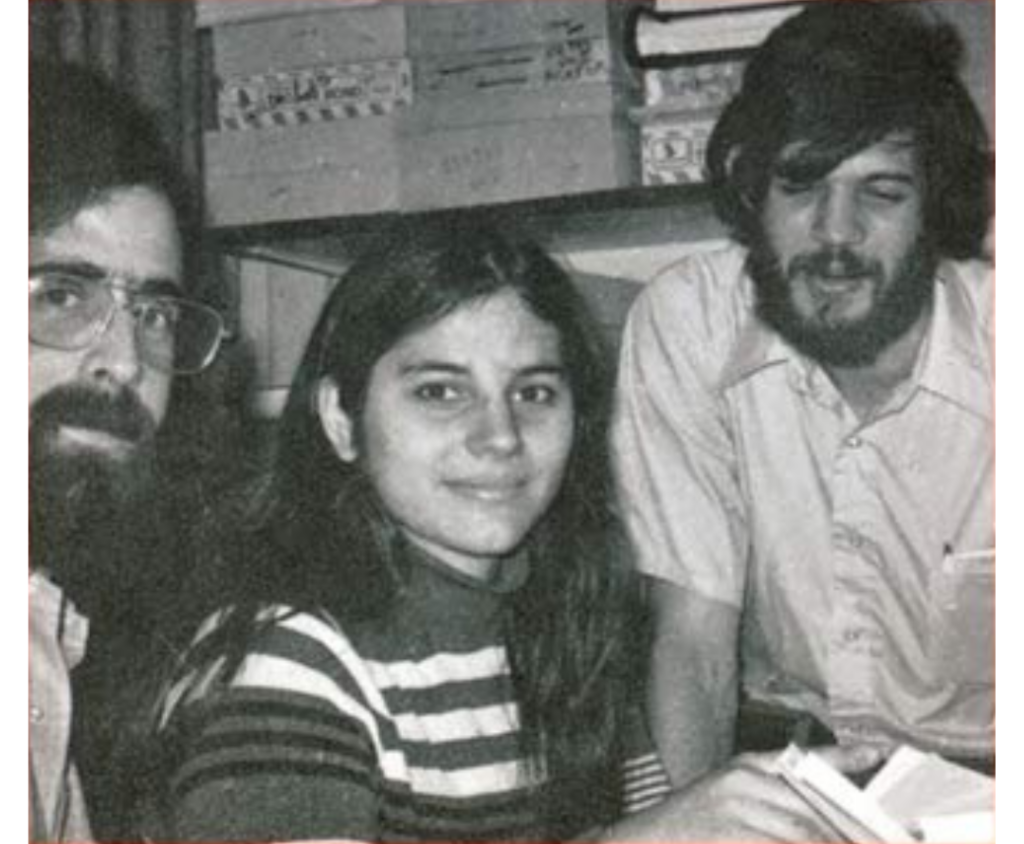 Three people sitting together, smiling, with books and boxes in the background. Black and white photo.