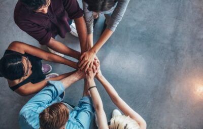 A group of people standing in a circle with their hands joined in the center over a gray floor.