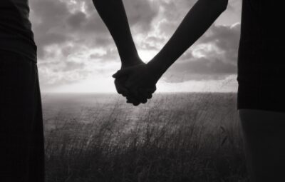 Two people holding hands in front of a cloudy horizon, with tall grass in the foreground.