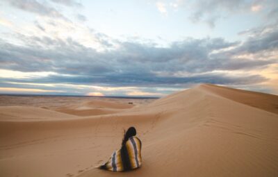 Person wrapped in a blanket sitting on a sand dune under a cloudy sky at sunset.