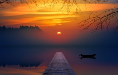 Sunset over a calm lake with a wooden pier and silhouetted boat, framed by tree branches and a misty horizon.