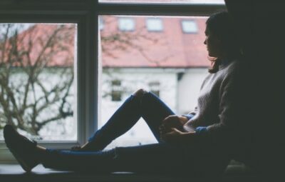 Person sitting by a window, looking outside at trees and rooftops on a cloudy day.