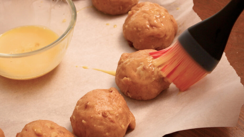Brushing egg wash on dough balls on parchment paper, with a glass bowl nearby.