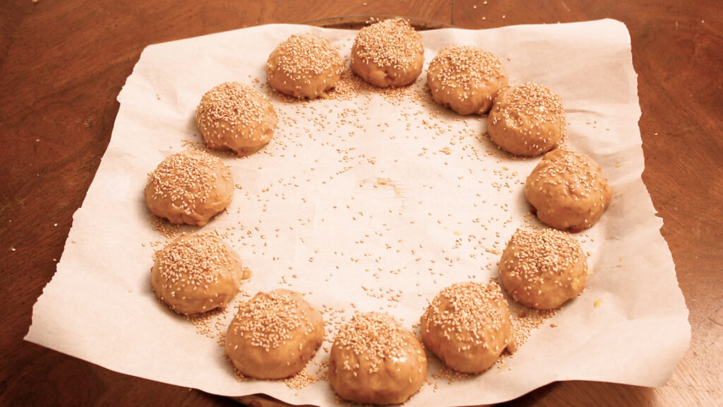 Twelve sesame seed-topped dough balls arranged in a circle on parchment paper.
