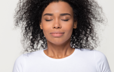 Person with curly hair and closed eyes, wearing a white shirt, looking serene against a plain background.
