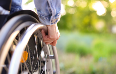 A person in a denim jacket using a wheelchair outdoors with a blurred green background.