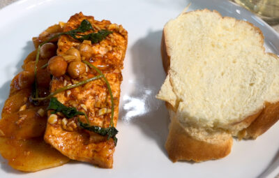 A plate with grilled chicken topped with herbs and chickpeas, potato slices, and a slice of bread.