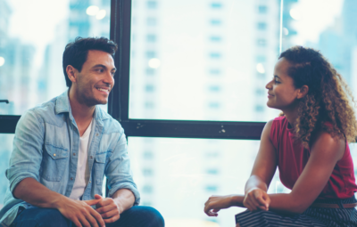 A man and woman sitting and talking by a window with a cityscape background.