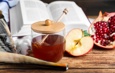 Honey jar, apple slices, pomegranate, open book, and shofar on a wooden table.