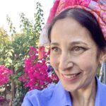 A woman smiling outdoors with a colorful headscarf, vibrant pink flowers, and greenery in the background.