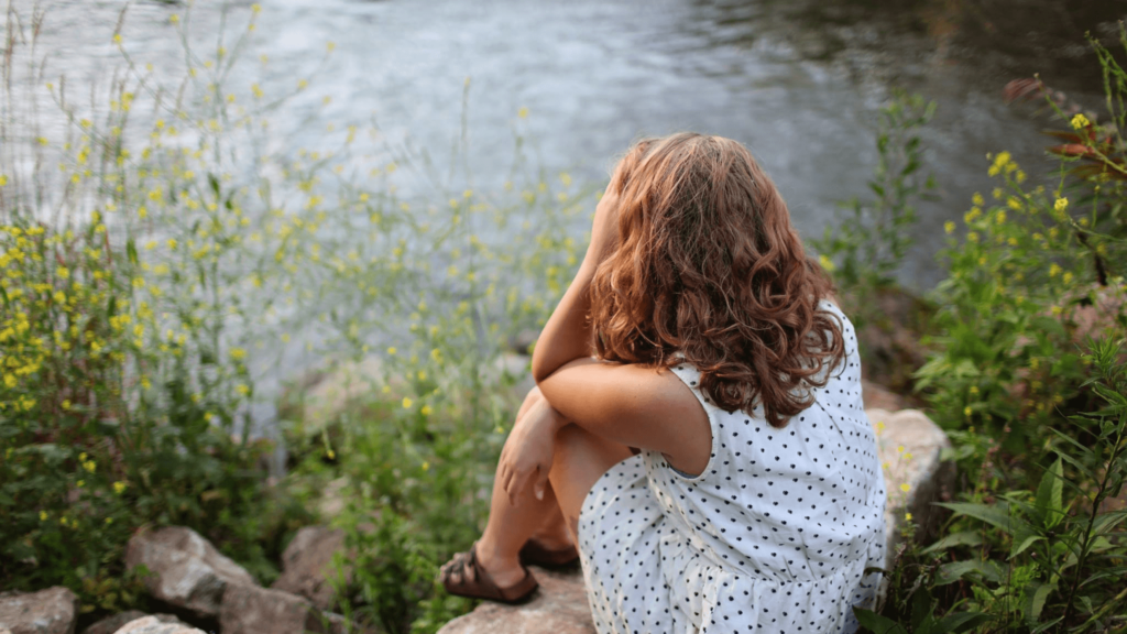 Woman with wavy hair wearing a black and white polka dot dress sits near a lake