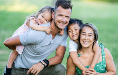 A happy family of four posing outdoors with smiles and piggyback rides in a grassy area.