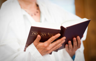 A person holds an open book with Hebrew text, wearing a white garment.