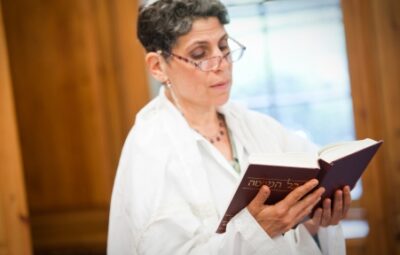 A person in a white robe reads from a book in a well-lit room.