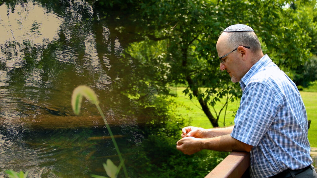 Man in a checkered shirt and kippah gazes thoughtfully over a bridge at a serene river surrounded by greenery.