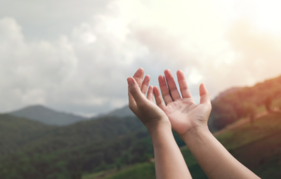 Hands reaching up towards a bright sky over a green mountainous landscape.