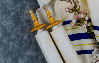 A Torah scroll with wooden handles next to a tallit and pink flowers on a textured surface.