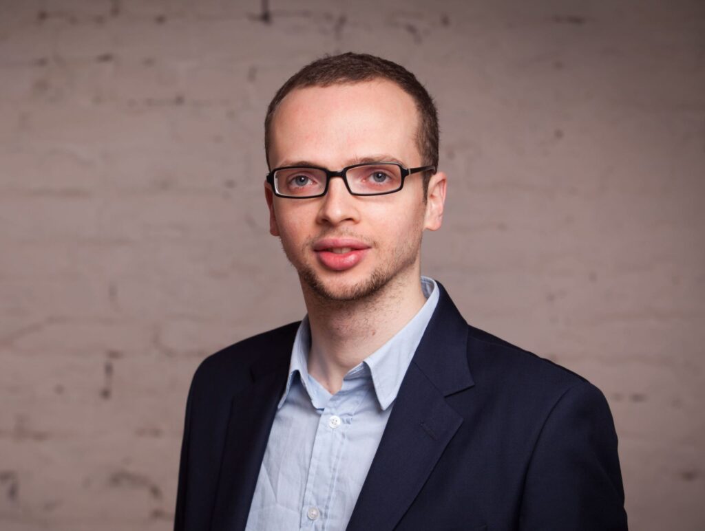 A person with glasses and short hair wearing a suit and light blue shirt, standing against a brick wall background.