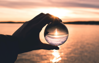 Hand holding a glass sphere with an inverted sunset reflection over a calm body of water.