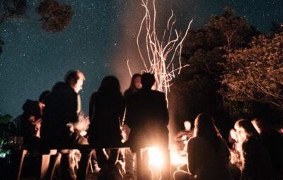A group of people sit around a campfire under a starry night sky, with trees silhouetted in the background.