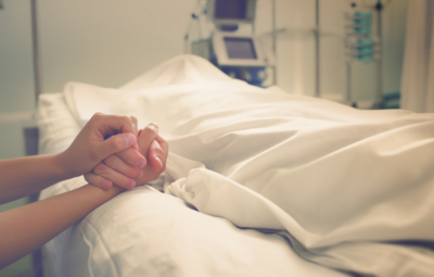 Person holding a hand of someone in a hospital bed, with medical equipment in the background.