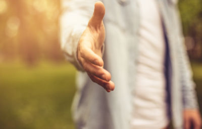 Person extending hand for a handshake, with a blurred outdoor background and warm lighting.