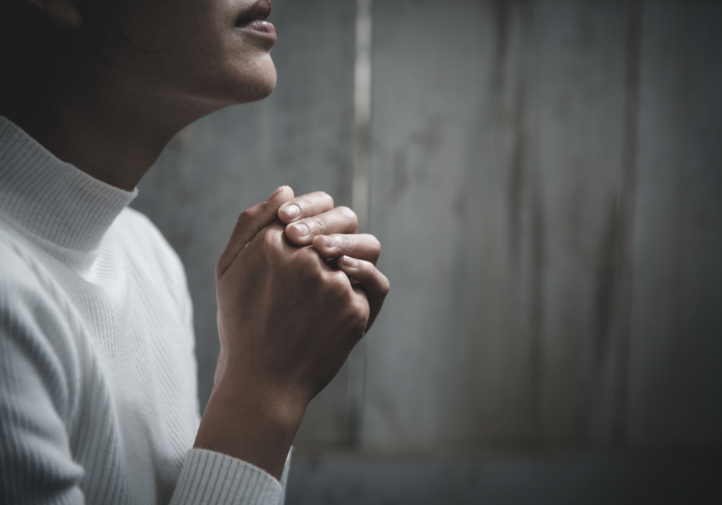 Person in a white sweater sitting with clasped hands, appearing contemplative or in prayer, against a wooden background.